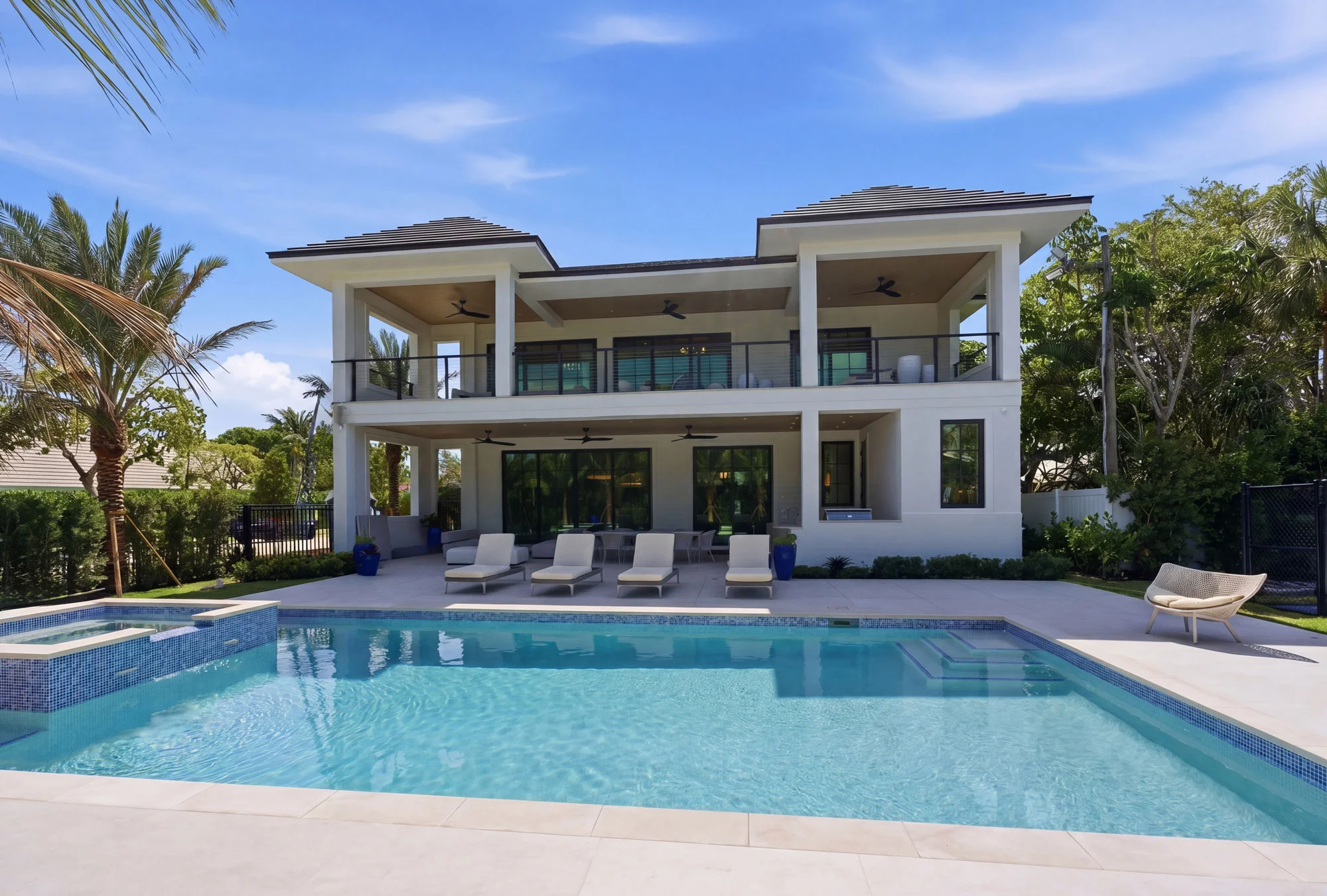 Modern two-story house with large windows and balconies, surrounded by lush greenery, features a spacious backyard with a swimming pool, lounge chairs, and a patio area under a clear blue sky.