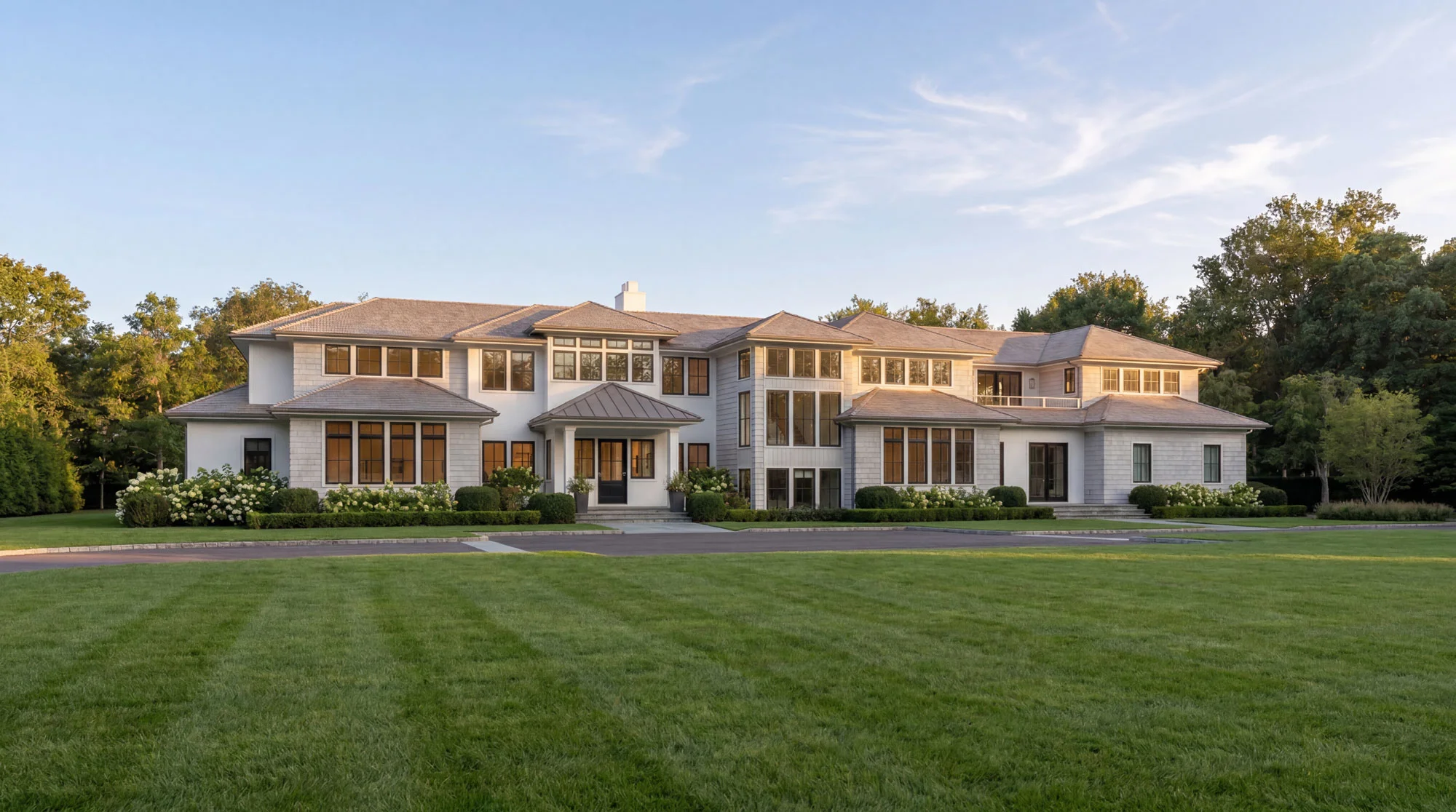Large, modern two-story house with many windows, a gray roof, and white exterior, surrounded by green lawn and trees under a clear blue sky.